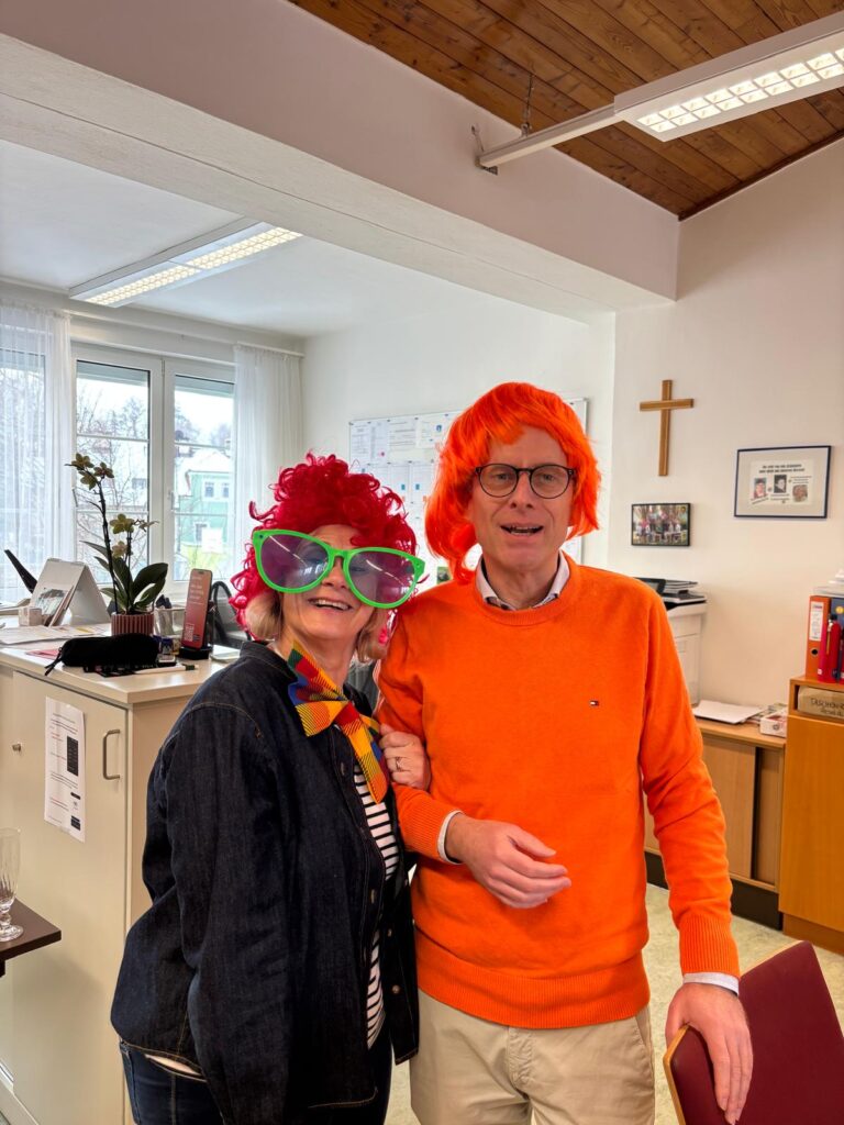 Frau Direktor mit roter Lockenperücke und übergroßer grüner Brille als Clown sowie ein Lehrer mit orangefarbenem Pullover sowie orangefarbener Perücke