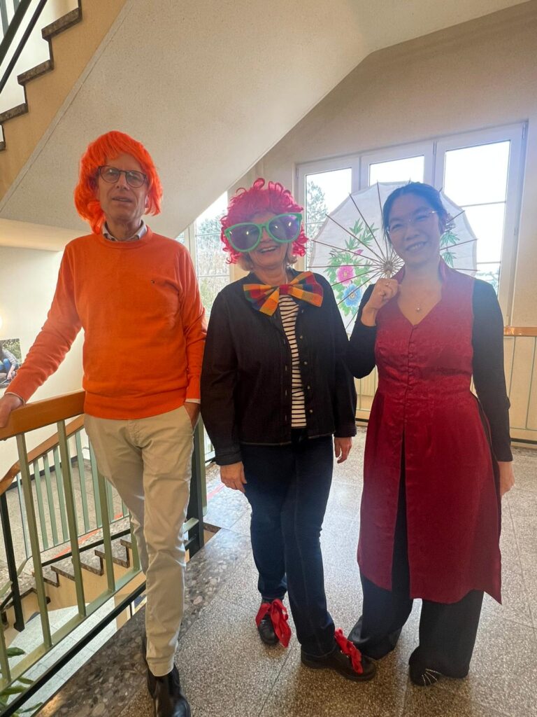 Frau Direktor mit roter Lockenperücke und übergroßer grüner Brille als Clown sowie ein Lehrer mit orangefarbenem Pullover sowie orangefarbener Perücke und eine Lehrerin mit schwarzen Haaren in einem asiatischem Kleid und weißem Schirm mit Blumendruck