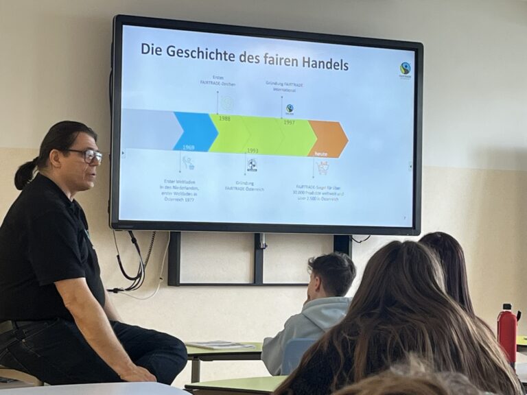 Der Vortragende sitzt schräg vor der digitalen Tafel mit einer Präsentation üder die Geschichte des fairen Handels