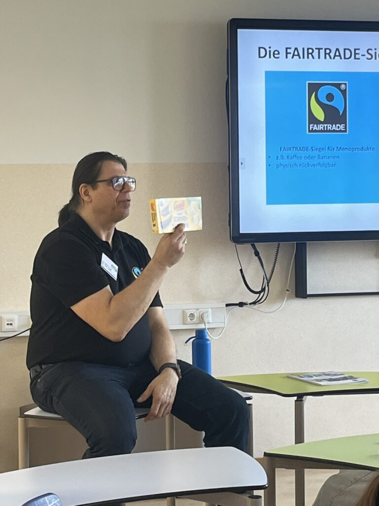 Der Vortragende sitzt auf einem Tisch und hält eine Packung Fairtrade Schoko-Bananen hoch