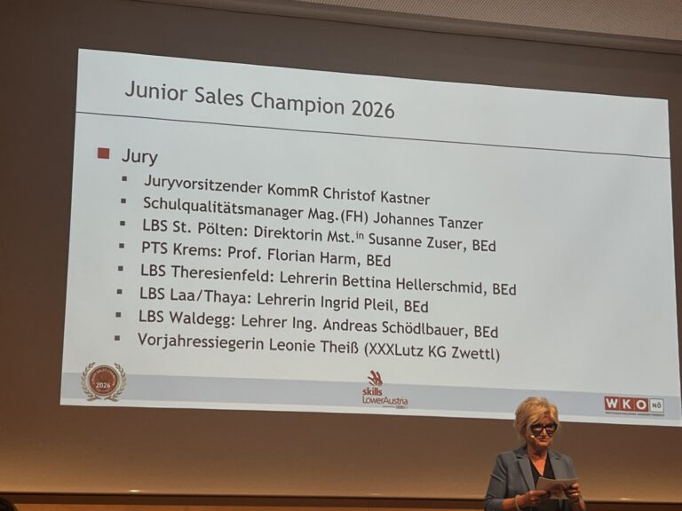 Beamerprojektion an der Wand mit acht namentlich genannten Jurymitgliedern des Junior Sales Champion 2026, davor die Moderatorin des Events mit kurzen blonden Haaren sowie schwarzer Brille