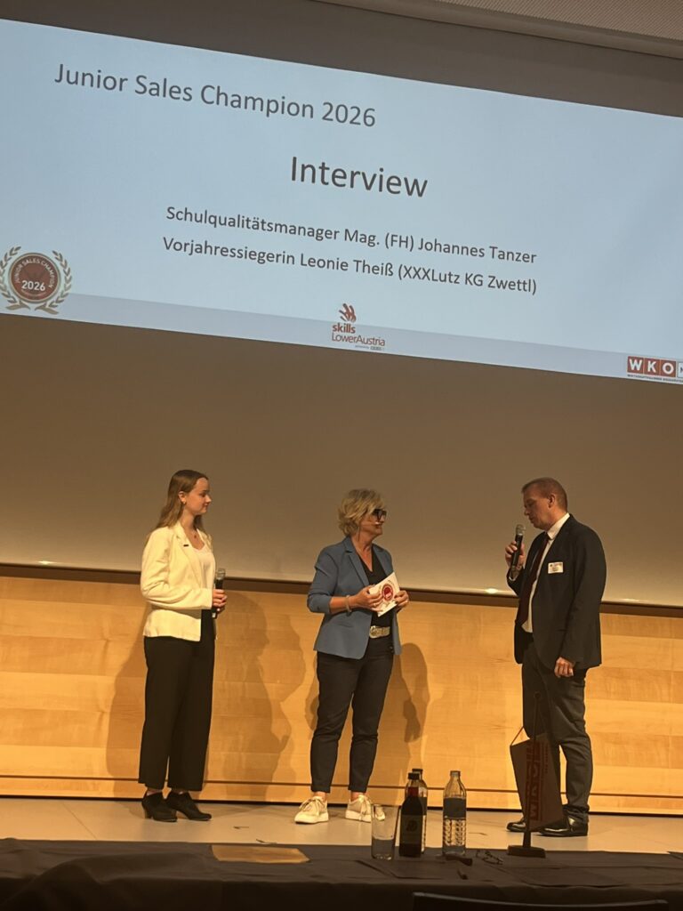 Moderatorin des Events mit Schulqualitätsmanager Mag. Johannes Tanzer und Vorjahressiegerin Leonie Theiss vom XXXLutz KG Zwettl im Interview auf der Bühne, dahinter eine Beamerprojektion an der Wand mit deren Namen