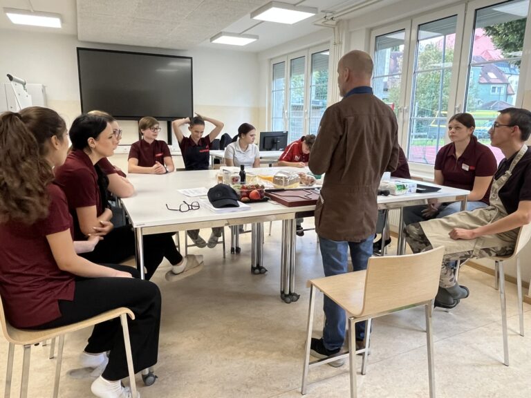 Klassenzimmer mit einem großen quadratischen Tisch, an dem Schülerinnen und Schüler sitzen mit einem Herrn mit Glatze in Rückansicht als Vortragenden