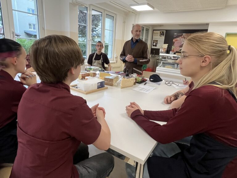Schülerinnen und Schüler sitzen am Tisch, auf dem verschiedene Käsestücke zu sehen sind, und hören einem Herrn als Vortragenden zu