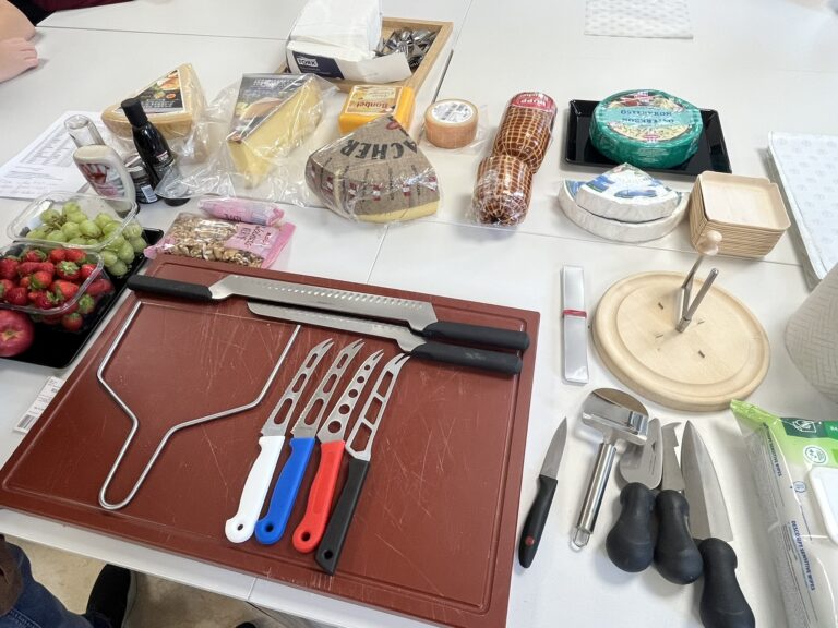 Arbeitsplatz mit verschiedenen Käsesorten, Erdbeeren, Weintrauben, Nüssen, Schneidebrett, verschiedene Käseschneidemesser, Käsehobel und Girolle