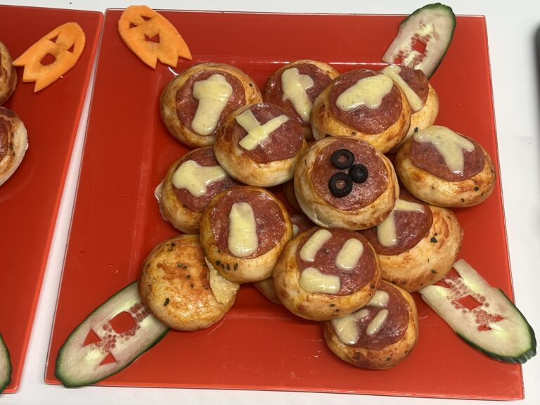 Platte mit Mini-Pizzen mit Salami, Oliven und Käse. Als Dekoration wurden dünne Scheiben aus Karotten und Gurken in Geisterform geschnitten.